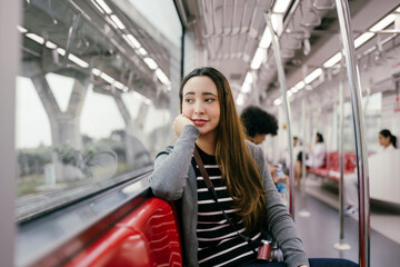 Young woman tourist with camera is sitting on a train, resting her head on her hand and looking out the window while traveling to her destination