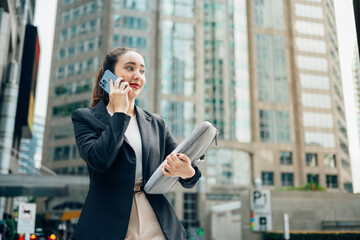 Businesswoman walking through a bustling city, holding a laptop case while engaged in a phone...