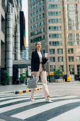 Young manager walking confidently across a pedestrian crossing, carrying a laptop bag while sipping takeaway coffee. Modern office buildings and skyscrapers create a vibrant urban backdrop