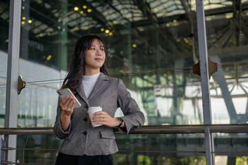 Young businesswoman is holding smartphone and coffee cup while waiting for her flight at airport terminal, she's wearing gray jacket and white turtleneck shirt