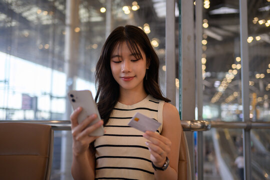 Asian woman holding a mobile phone and credit card, engaging in online shopping while waiting for her flight at the airport terminal, blending travel and modern technology seamlessly