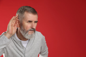 Senior man showing hand to ear gesture on red background, space for text