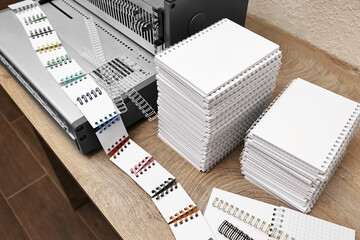 Modern binding machine, stacks of notebooks and double loop wire binding spines on wooden table indoors