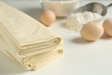 Stack of raw puff pastry dough, eggs and flour on white table, closeup