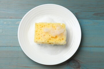 Yellow sponge with foam and plate on light blue wooden table, top view
