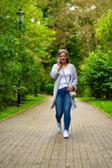 Fototapeta premium Beautiful middle aged woman walking and talking on phone against background of greenery in city park. Mobile phone conversation in park. Front view