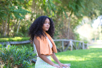 Young, beautiful, Latin, brunette woman with curly hair, dressed in sports clothes and wearing a towel around her neck to dry her sweat, is sitting on a wooden fence. Concept of sport and health.