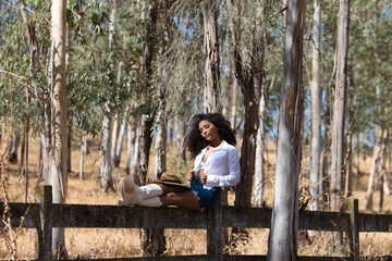 Fototapeta premium woman, young, beautiful, Latin, brunette with curly hair dressed in white shirt, denim skirt, white boots and hat, sitting with her legs stretched out on the forest fence. The woman rests.