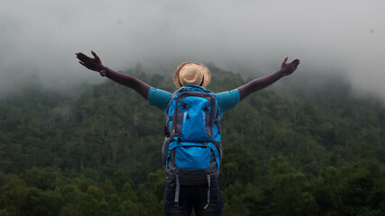 African hiker with hat and backpack stands with his arms outstretched on top of a mountain. Concept of traveling and hiking adventures.