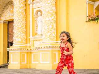 Beautiful girl in red clothes runs in front of the yellow Catholic church with white details. Colonial church in Antigua Guatemala.