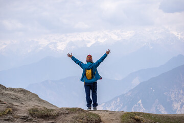 A woman stands triumphantly atop a mountain peak with arms wide open, wearing a blue jacket, yellow backpack, and colorful woolen cap. Snow capped Himalayan mountains and cloudy skies in the backdrop.