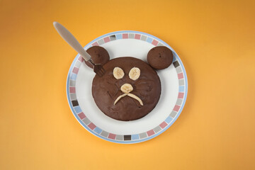 Homemade chocolate cake with eyes, nose and mouth made from banana. Sad face of brown bear with fork stuck in it. Chocolate cupcakes on plate in shape of bear head