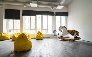 Empty kindergarten room with white walls, big windows, rocking horse and bunch of yellow bean bag...