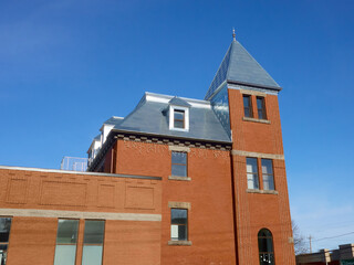 A red brick building features a distinctive tall tower with a pointed roof. The structure displays various windows and a decorative roof, set against a clear blue sky. 