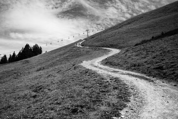 A winding road ascends through grassy hills under a cloudy sky. Hikers can be seen making their way...
