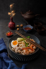 Udon noodles with egg, shrimp, mushroom, red hot chilli peppers, on a dark rustic background with tea cups and dry flower in a red vase