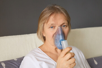 A woman sits comfortably at home, using a nebulizer device for respiratory treatment. Natural light...