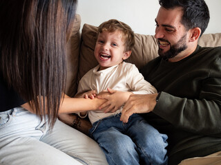 A young boy laughs joyfully while surrounded by his parents on a comfortable couch. The family enjoys playful moments together, creating a warm and loving atmosphere in their home.