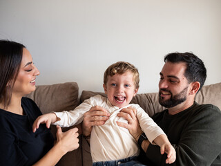 A cheerful young boy laughs while being playfully held by his parents on a cozy couch. The warm...
