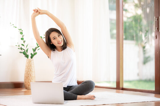 Happy smiling Asian woman doing stretching warm up exercise in living room at her house with laptop on the floor
