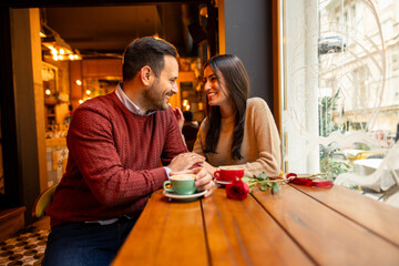A happy Caucasian couple, a mid adult man and woman, enjoy a warm coffee date in a cozy cafe setting. Sharing smiles and conversation, with roses and cups on the wooden table beside them.