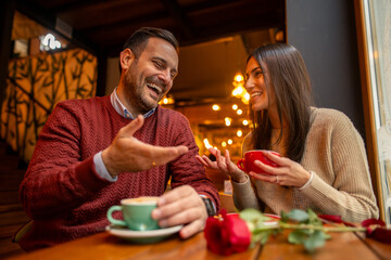 A happy Caucasian mid adult couple celebrates International Women's Day at a cozy cafe. The man, in a red sweater, and the woman, in a beige sweater, enjoy hot drinks and a rose.