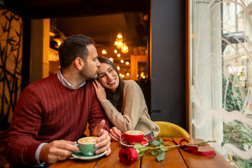 A happy Caucasian mid-adult couple, with one partner a barista and the other a teacher, enjoys coffee in a cozy cafe. Both are in casual attire, surrounded by warm lighting and a rose on the table.