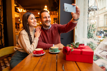A cheerful Caucasian mid adult couple, dressed in casual sweaters, enjoys taking a selfie in a cozy cafe. The setting features warm lighting, coffee cups, and a romantic gift on Valentine's Day.