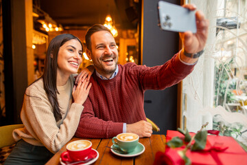 A cheerful Caucasian mid adult couple, dressed in casual sweaters, enjoys taking a selfie in a cozy cafe. The setting features warm lighting, coffee cups, and a romantic gift on Valentine's Day.