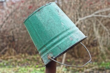 one enameled round old industrial dirty garden with handle empty green bucket hanging on rusty iron pole during daytime outdoors