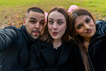 Fototapeta premium three young male and female friends take a selfie having fun sitting on a bench in a public park in winter