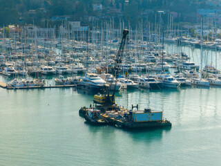 Crane barge in marina with many moored yachts and sailboats.