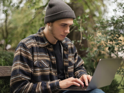 un joven moderno con est&eacute;tica hipster. Lleva un gorro y una camisa de franela y trabaja desde un parque con una computadora port&aacute;til
