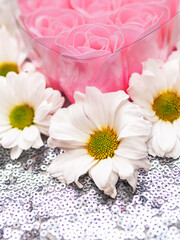 A heart made of six pink roses. The roses are surrounded by white daisies and places on silver glitter background. Concept of love and affection. Abstract conceptual art.