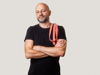 Bald male with slim athletic body wearing black shirt and holding red color resistance band. Man is in his 50s with grey short beard. Fitness couch with friendly face and attitude. Light background.