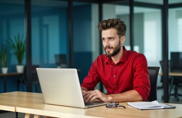 Fototapeta premium Young male programmer works on laptop in office. Sits at desk, looking thoughtfully at monitor, video call. Wears red shirt. Modern office environment. Work, tech, creative, design, tech, success,