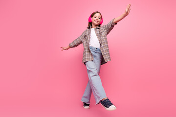 Obraz premium Young girl smiling and posing against pink background while enjoying music with headphones in a trendy casual outfit.
