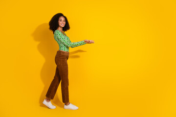Cheerful young woman in green top and brown pants posing against yellow background with inviting gesture