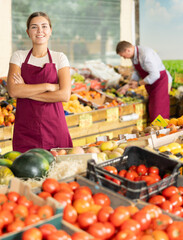 Portrait of confident smiling young salesgirl in maroon apron standing with arms crossed among produce stands with ripe fresh organic fruits and vegetables in greengrocery....