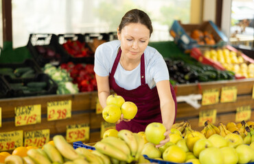 With practiced hand, woman supermarket worker arrange apples in neat pyramid by window. Female seller of vegetable department of store is replenishing showcase with apple, puts ones on showcase