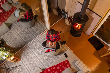 Cozy cabin workspace with a laptop and fireplace. Overhead view of a woman in a red plaid shirt working on a laptop in a cozy cabin, sitting on a pouf by a warm fireplace.