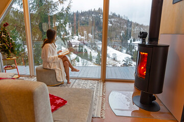 Relaxing by the fireplace with a snowy mountain view. A woman in a white robe sits on a pouf...