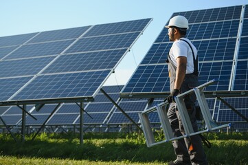 Portrait of Young indian man technician wearing white hard hat near solar panels against blue sky....