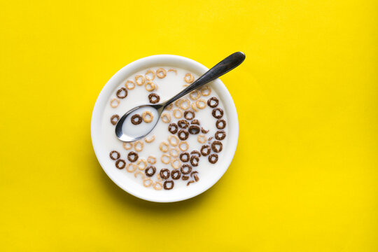 A mix of chocolate and plain cheerios floating in milk in a plain white bowl. A spoon floats in the center of the bowl