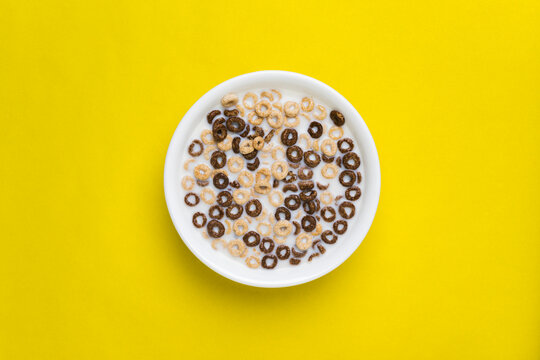 A mix of chocolate and plain cheerios floating in milk in a plain white bowl on a yellow background