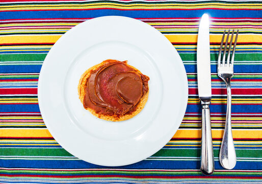 A pancake covered in dulce de leche on a large white plate with a silver knife and fork on a multicolored placemat