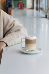 A cozy café scene featuring a layered latte macchiato in a clear glass mug on a white saucer. A person wearing a beige sweater sits at the table, adding warmth to the minimalist, modern setting.