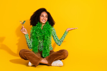 Charming young woman with curly hair and green outfit posing against a vibrant yellow background holding a cocktail glass