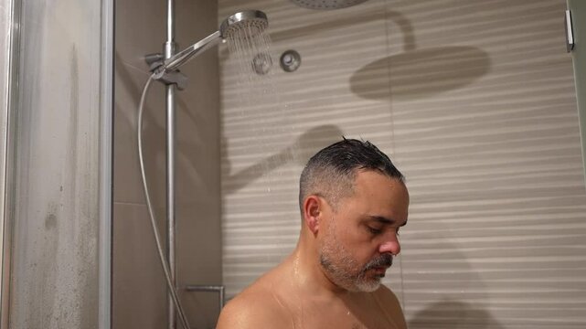 Male washing body under warm water stream, lathering skin with soap while enjoying relaxing shower moment in home bathroom