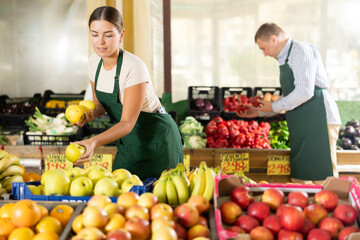 Young woman seller of garden stuff department store is replenishing showcase with fruits, puts...